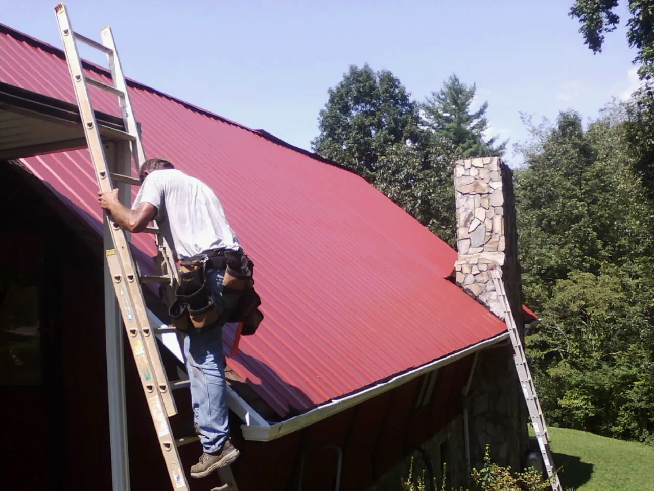 Roofer on a ladder beside a red metal roof and stone chimney — Soffit & Fascia Repair by Roofing Brothers in Congers